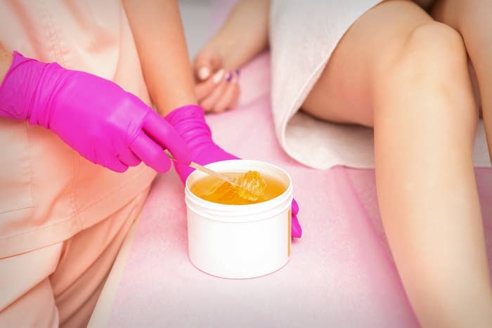 Person med hansker som forbereder voks for hårfjerning. En klient ligger i nærheten med bena delvis dekket