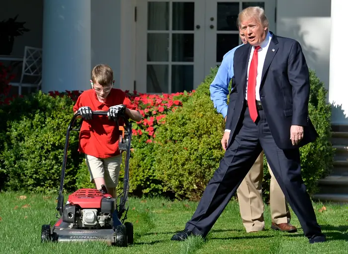 Et barn klipper plenen i Det hvite hus mens en voksen i dress med rødt slips observerer og gestikulerer animert