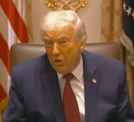 En mann i dress taler på et formelt arrangement med flagg i bakgrunnen