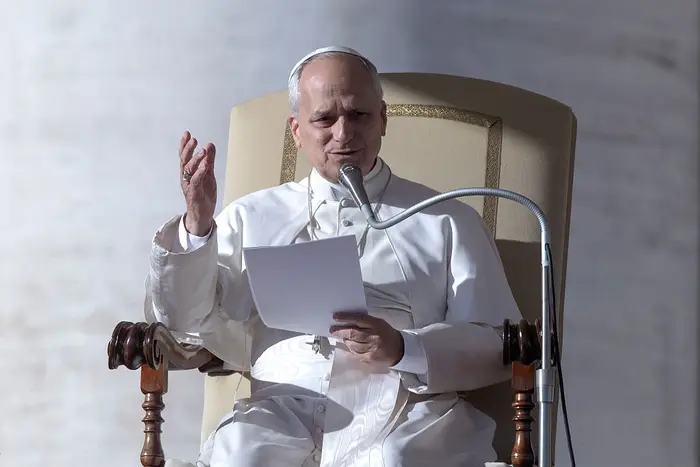 En person i pavelig antrekk snakker i en mikrofon mens han holder et papir, sittende på en stor stol under et arrangement