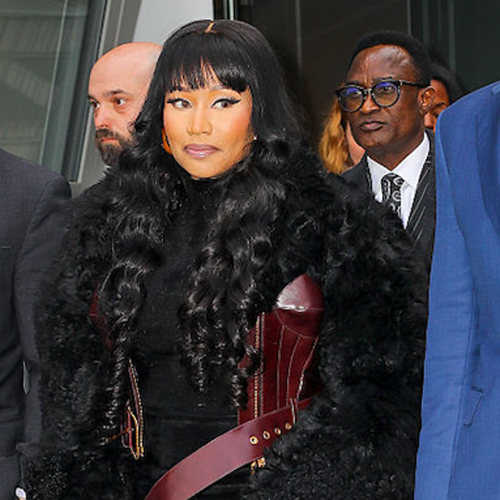 Person i svart pels og burgunderbelte går, holder en veske. Andre i formell antrekk i nærheten, utendørs