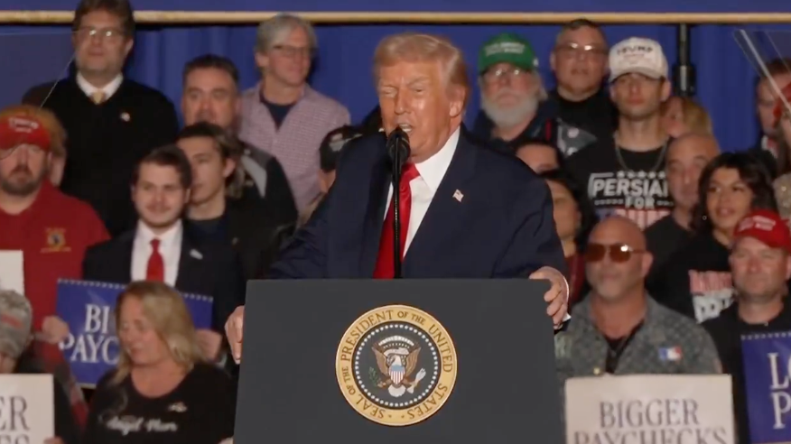 En person snakker på et podium med en gruppe mennesker bak. På skilt i mengden står det «Større lønnsslipp». Scenen ser ut til å være på et arrangement eller et rally