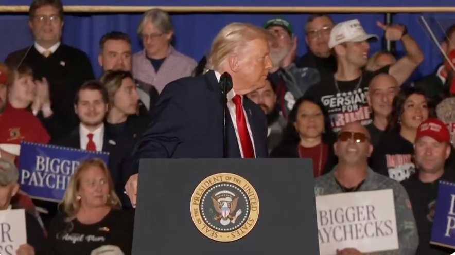 En person på et podium med en presidentsegl henvender seg til en folkemengde. Folk holder skilt med teksten 