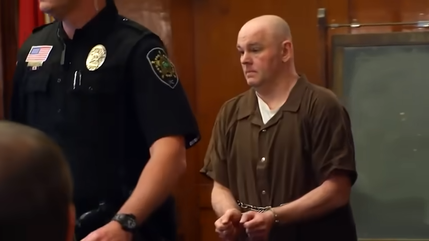 En person i en rettssal, iført en brun fengselsdress og håndjern, går med en politimann i nærheten