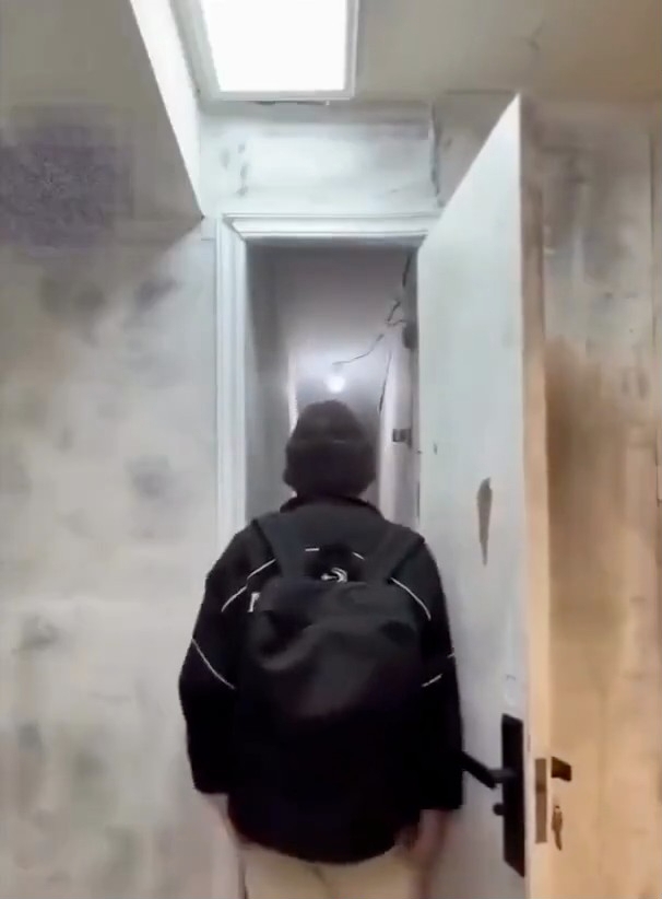 Person med ryggsekk som går inn i en smal, uferdig gang med synlige rør og ledninger i taket