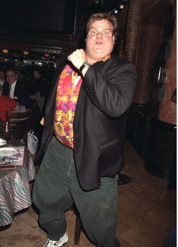 Person i en livlig, mønstret skjorte, jakke og løse bukser, poserer energisk på et arrangement, og virker leken og animert