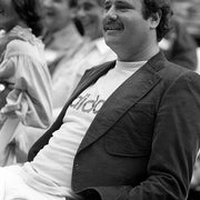 Person som sitter på et utendørsarrangement, iført en blazer over en skjorte med en synlig logo, smiler mens han ser fremover