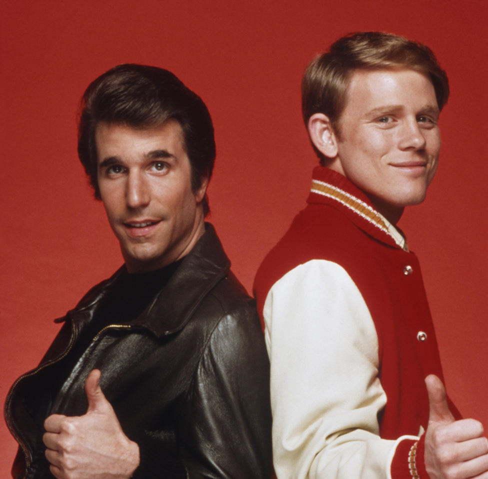 To personer med tommelen opp, den ene i en skinnjakke og den andre i en letterman-jakke, stående rygg-mot-rygg mot en vanlig bakgrunn