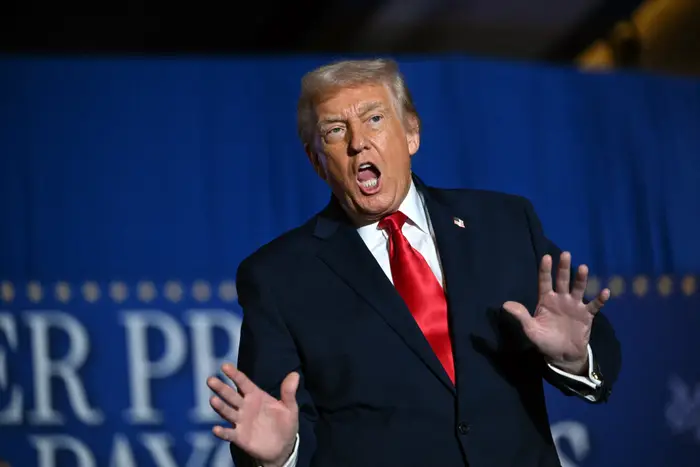 En person i mørk dress og rødt slips bevegelser med hendene hevet, ser ut til å snakke lidenskapelig på et arrangement