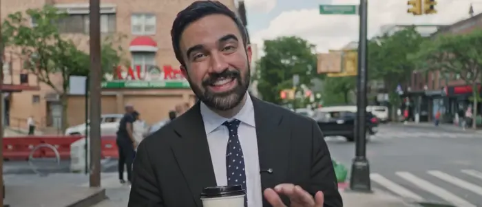 Person i dress som holder en kaffekopp og smiler i en bygate med bygninger og mennesker i bakgrunnen