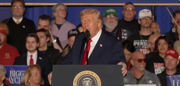 En person taler ved et podium med en presidentsegl, omgitt av en folkemengde ved et offentlig arrangement