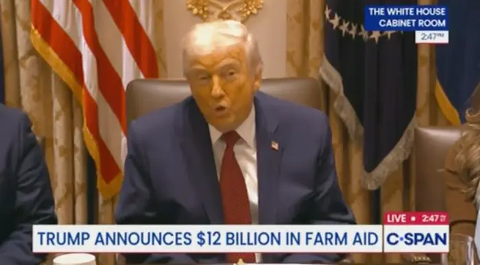 En mann snakker på et møte i et rom med amerikanske flagg. På banneret står det: 