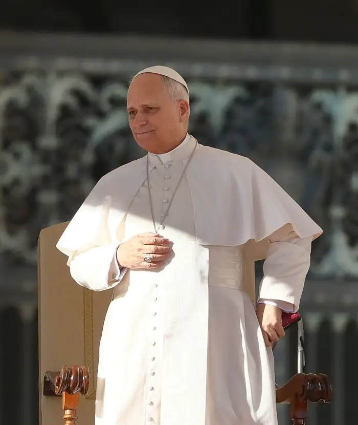 En religiøs leder i tradisjonell antrekk står utendørs, holder en hånd på en utsmykket stol, og deltar i et offentlig arrangement