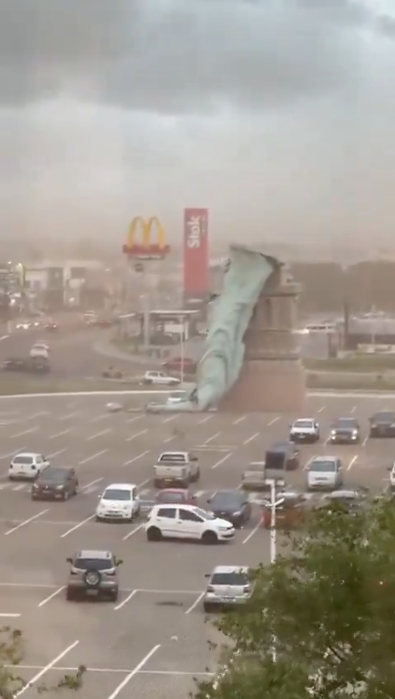 Kollapsert Frihetsgudinnen kopi liggende over en parkeringsplass, omgitt av biler, med gatekjøkkenrestaurantskilt i bakgrunnen