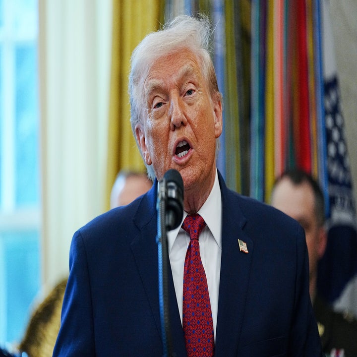 En mann i dress snakker ved en mikrofon i en formell setting med fargerike dekorasjoner i bakgrunnen