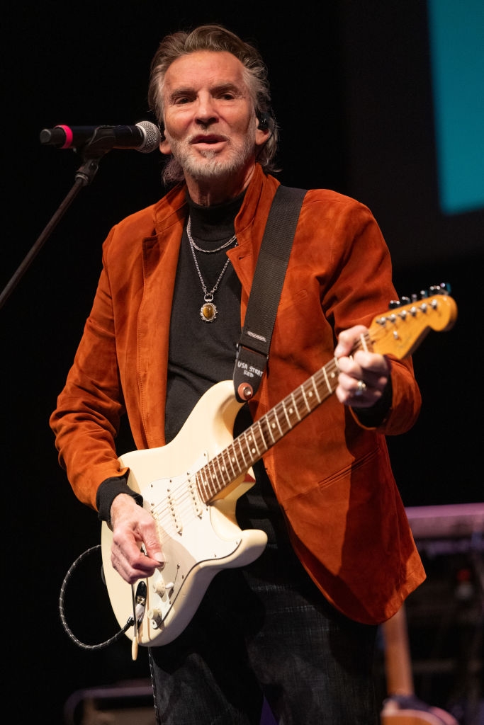 Musiker som opptrer på scenen med en gitar, iført en stilig jakke og lagdelte halskjeder, holder en mikrofon med et fokusert uttrykk