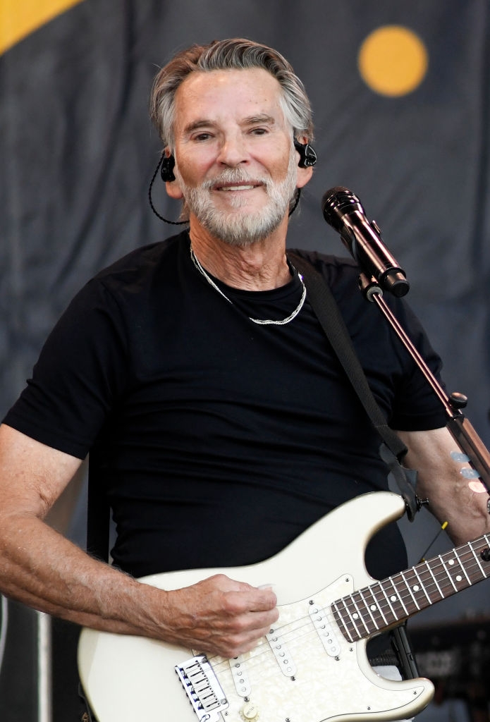 Musiker på scenen holder en gitar, iført en svart T-skjorte og hodesettmikrofon, opptrer foran et levende publikum