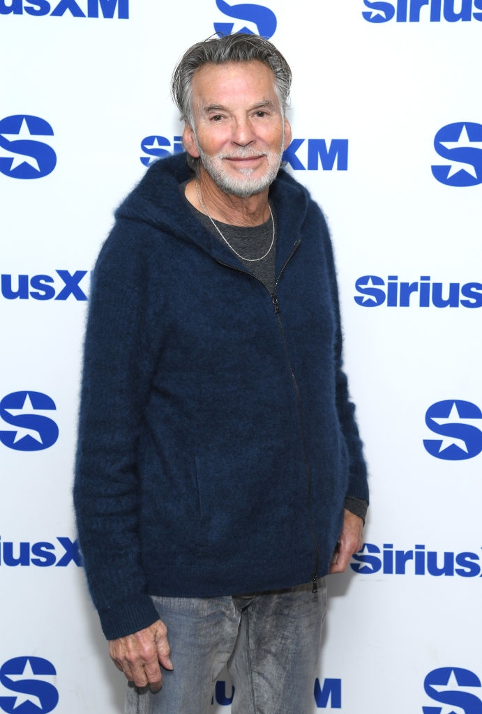 En mann i en uformell mørk cardigan og grå jeans, poserer foran et SiriusXM-bakteppe på en mediebegivenhet