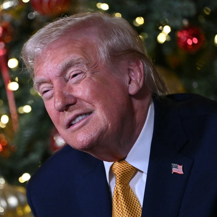 Person i formell antrekk smiler litt foran et pyntet juletre med lys og pynt