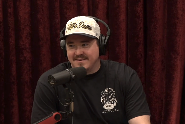 Person med hodetelefoner og Notre Dame-hette snakker inn i en mikrofon på en podcast mot et gardinbakgrunn