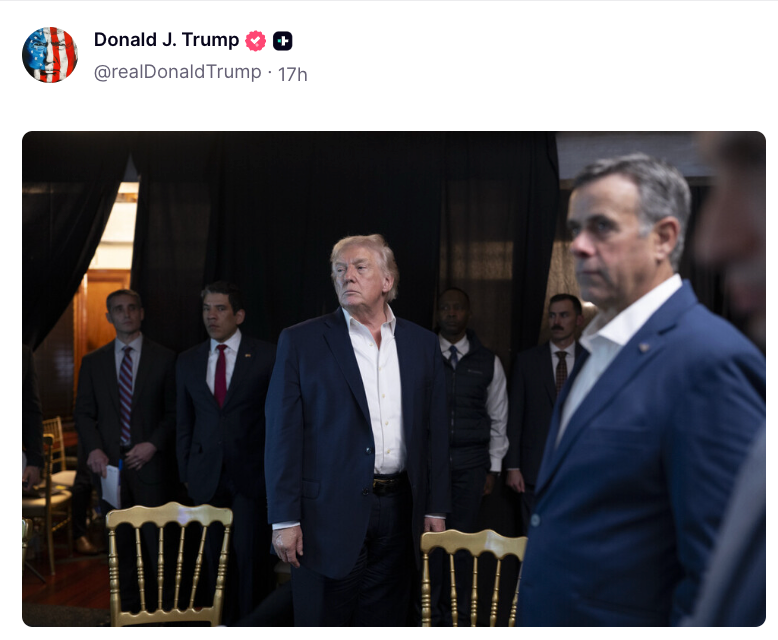 En gruppe menn i dress står i et svakt opplyst rom. Sentral skikkelse som ser fremover, flankert av andre, med oppmerksomme uttrykk