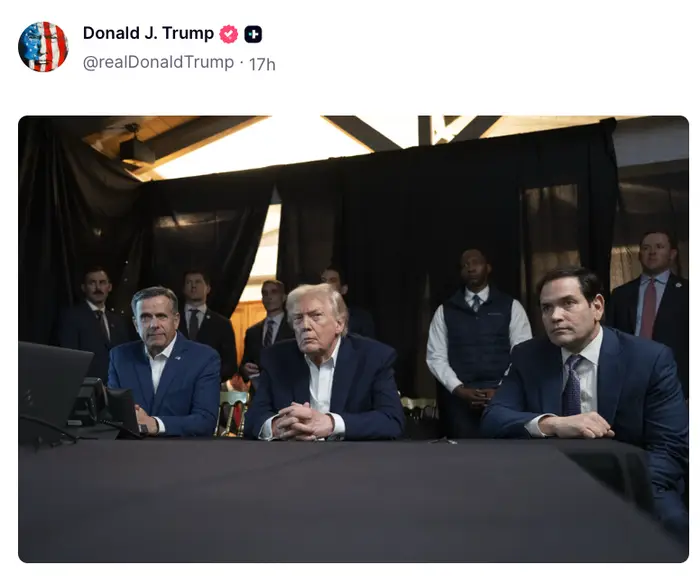 Tre dresskledde menn sitter bak et bord med alvorlige uttrykk, omgitt av andre i bakgrunnen