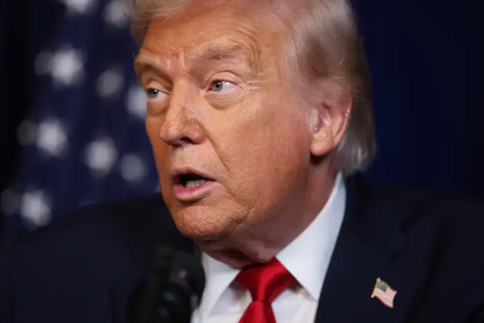 En person i dress og rødt slips snakker foran et bakteppe med et mønster, under et offentlig arrangement