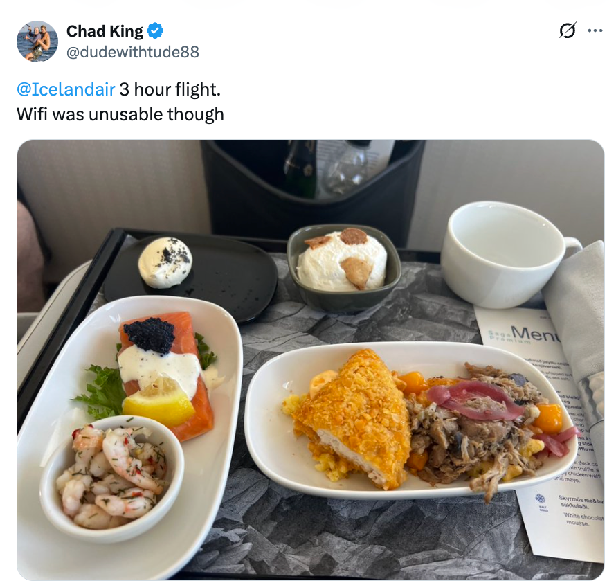 Flymåltid med reker, laks, en rett med løk og kjøtt, brød og dessert på et brett. Kopp og meny er synlig i bakgrunnen