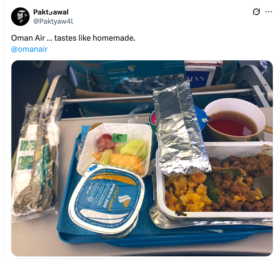 Flymåltid på et brettbord med frukt, innpakket redskaper og en kopp te, servert på en Oman Air-flyvning