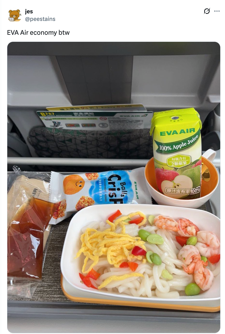 Flyselskapsmåltid med nudler, reker, edamame, egg i skiver, paprika, en matbit, rundstykke og eplejuicekartong på et flybrettbord