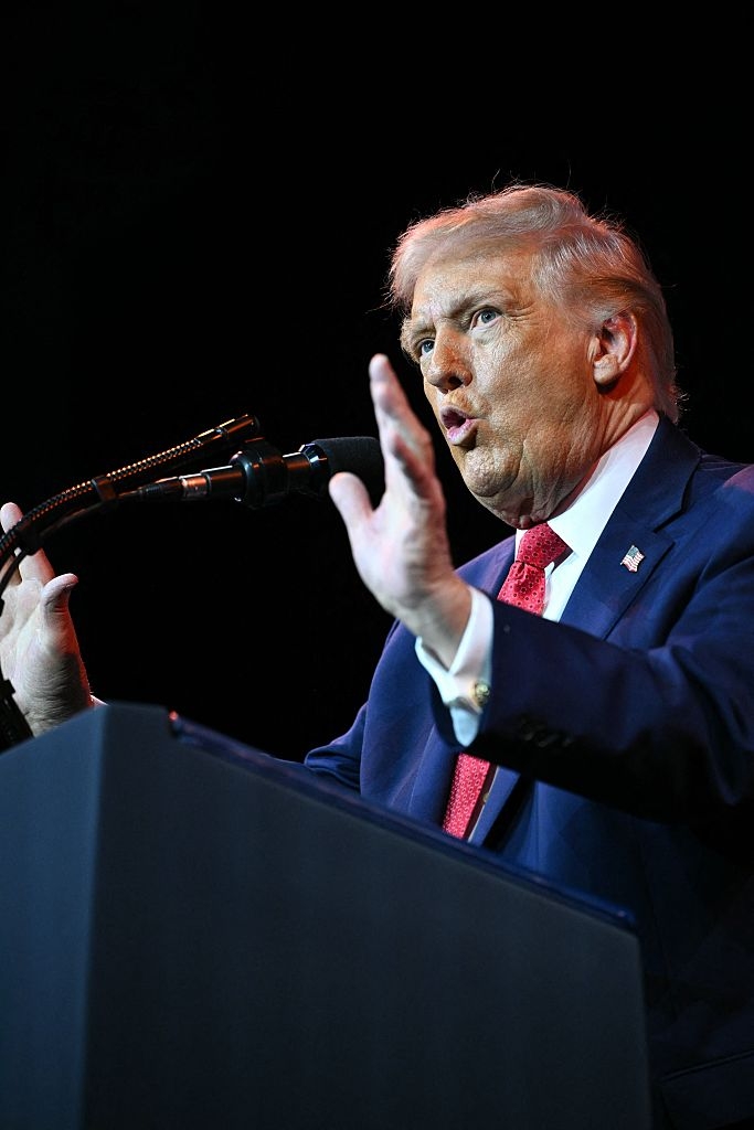 En person snakker animert på et podium, iført dress og rødt slips, på en scene med en mikrofon