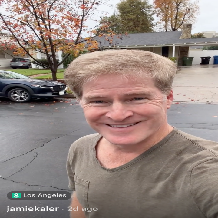 En person smiler mens han tar en selfie utendørs i en boliggate med biler og høsttrær synlige i bakgrunnen