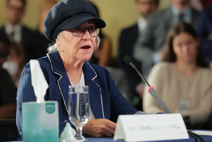 En eldre kvinne i blazer og hatt snakker inn i en mikrofon ved en formell begivenhet, sittende ved siden av et navneskilt med teksten