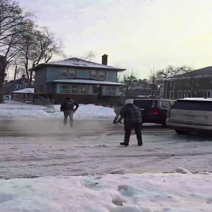 Folk som måker snø i en boliggate med to parkerte kjøretøy i nærheten og hus i bakgrunnen. Snø dekker bakken
