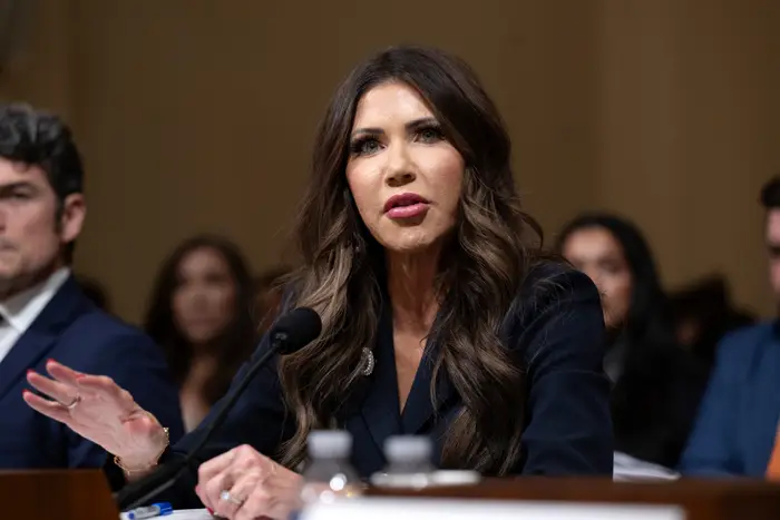 Person som snakker på en formell begivenhet, iført et mørkt profesjonelt antrekk, sitter ved et bord med mikrofoner foran seg. Andre personer er i bakgrunnen