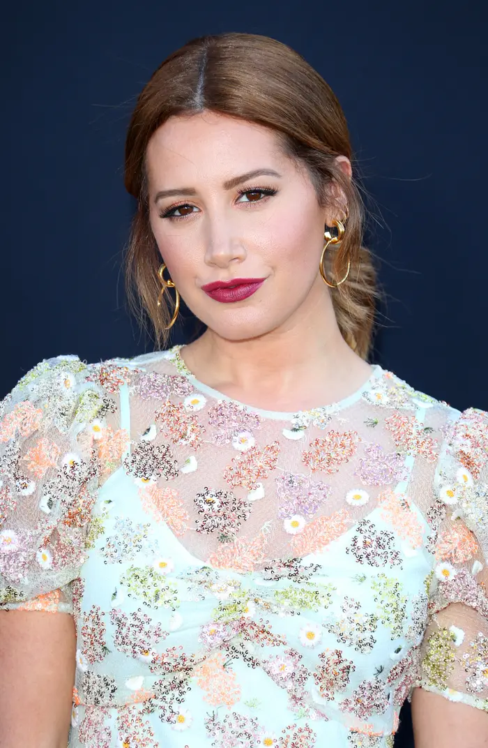 Ashley Tisdale i blomstermønstret kjole med puffede ermer og ringøredobber på mørk bakgrunn