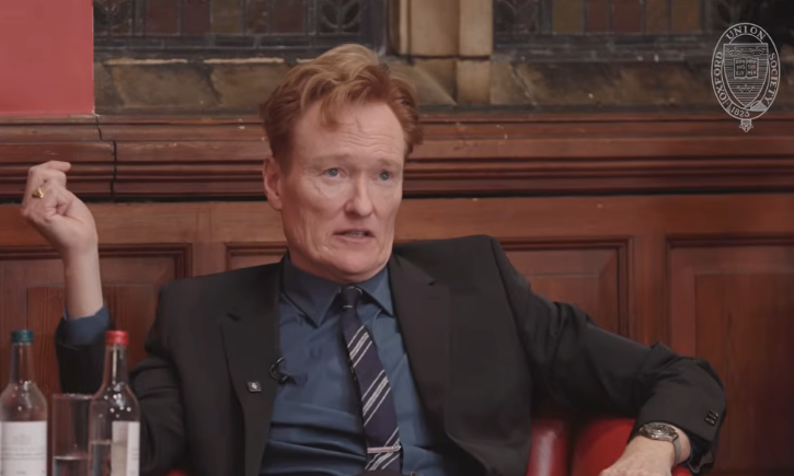 Person i dress med stripet slips snakker på en scene med et seriøst uttrykk, sittende mot et bakteppe av tre og stein