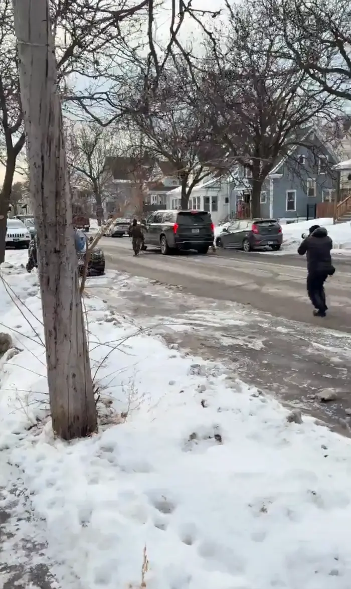 Snødekt gatebilde med folk som går og parkerte biler, vei omgitt av trær og hus i bakgrunnen. Fremstår rolig og beboelig