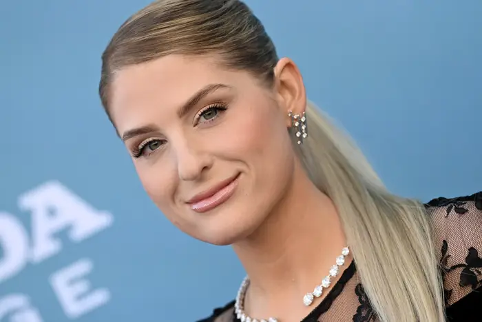 Meghan Trailor med slanke hestehale og juveløredobber poserer på den røde løperen, iført en ren blomsterkjole og et perlekjede mot et vanlig bakteppe