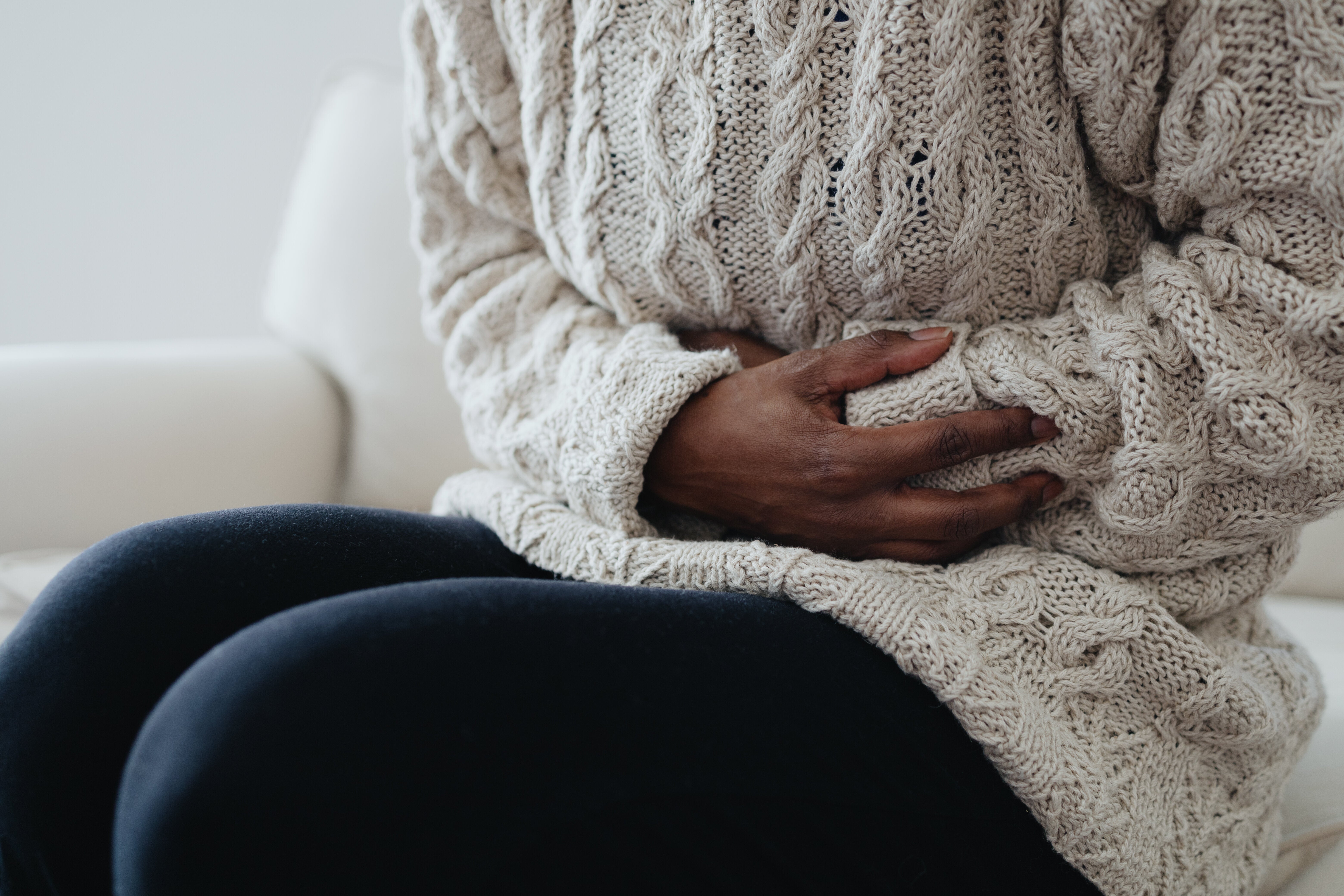 Person i strikket genser som sitter på en sofa, holder magen, opplever muligens ubehag