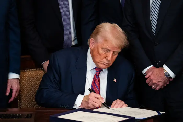 En mann i dress sitter ved et skrivebord og signerer et dokument, omgitt av andre i formell antrekk