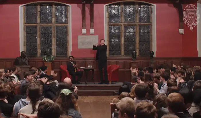 En foredragsholder henvender seg til et publikum i en storslått sal med glassmalerier, med statuer og ordene