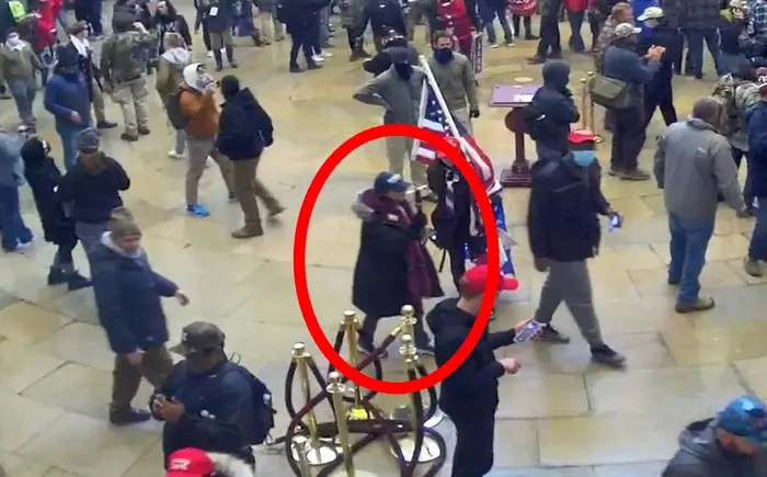 En folkemengde inne i en stor sal med flere mennesker gående og stående, noen iført hatter og masker. En person er ringt inn i rødt