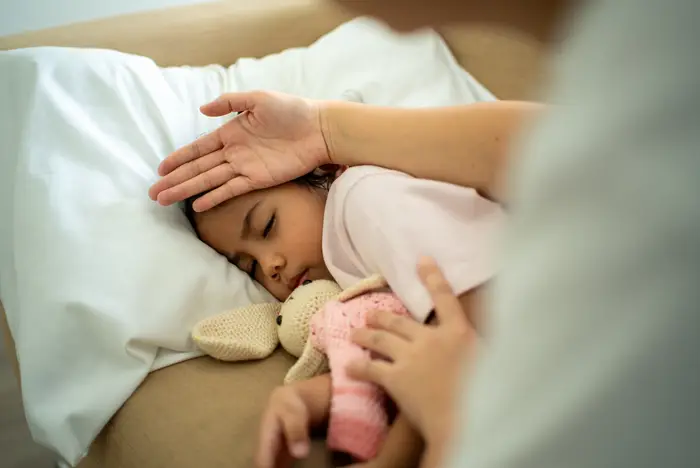 Barn som sover i sengen, holder en utstoppet leke. En voksen hånd sjekker forsiktig barnets panne