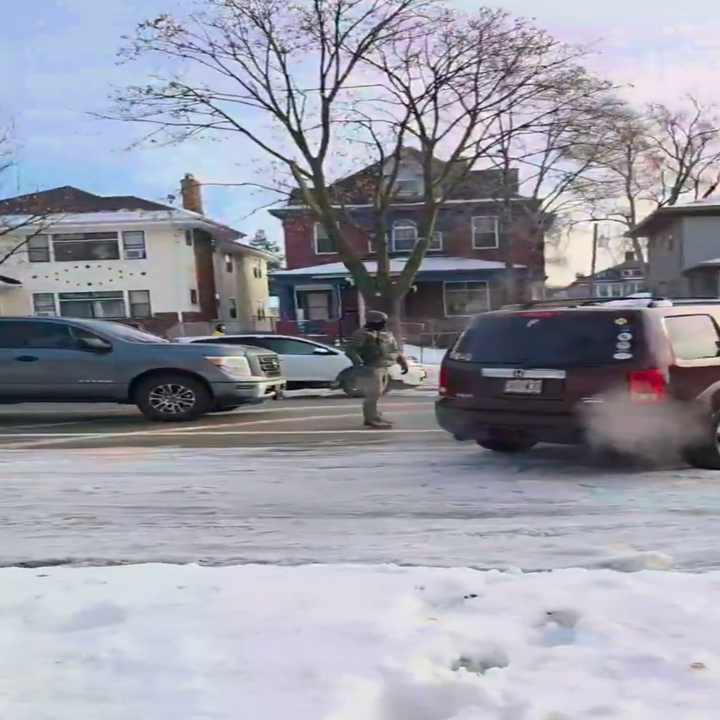 Person som går på snødekte forstadsgate med parkerte biler og hus; trær uten blader i bakgrunnen