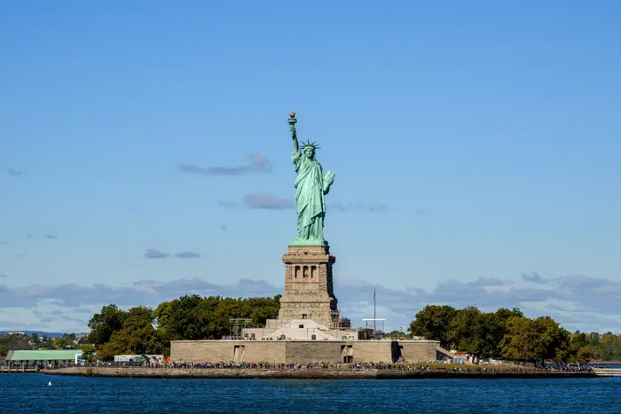 Frihetsgudinnen på Liberty Island, New York, omgitt av turister og trær, med klar himmel i bakgrunnen