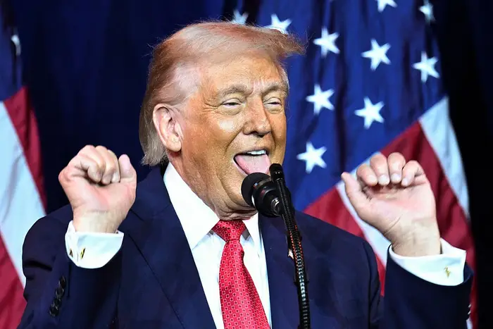 En person i dress og rødt slips bevegelser med begge hendene opp nær ansiktet bak en mikrofon, stående foran et bakteppe av flagg