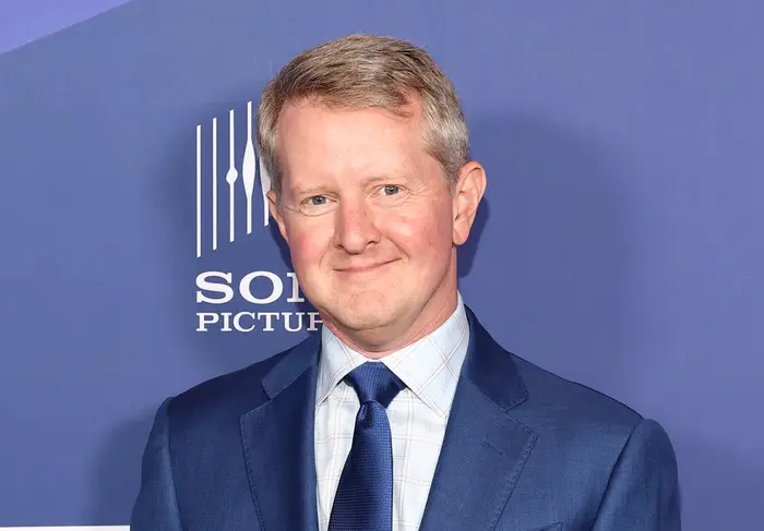 Ken Jennings i dress med slips ved et formelt arrangement, stående foran et bakteppe med logoer
