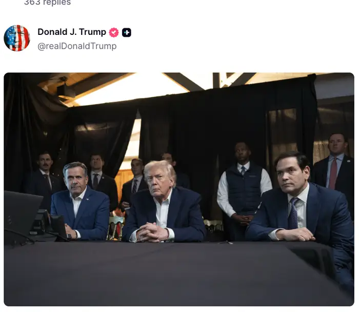 Donald Trump sitter ved et bord med to menn i dress, omgitt av folk i formell antrekk, under det som ser ut til å være et møte eller arrangement