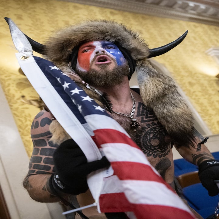 Person i hodeplagg med horn og ansiktsmaling som holder et flagg og megafon i innendørs omgivelser